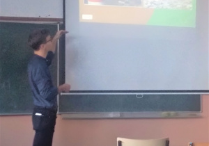 Prowadzący zajęcia stoi przy tablicy w sali lekcyjnej. Omawia slajd wyświetlany na ekranie.